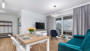 A modern living room with a wooden dining table, white chairs, a teal sofa, and a TV on the wall.