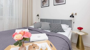 Una cama bien tendida con cabecera gris, una mesita de noche con lámpara, y una bandeja con comida y flores.
