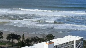A coastal view with waves crashing onto the shore, a building with a white facade and orange accents, and a clear sky. - Your Stay In Surfers (Surfers Paradise)