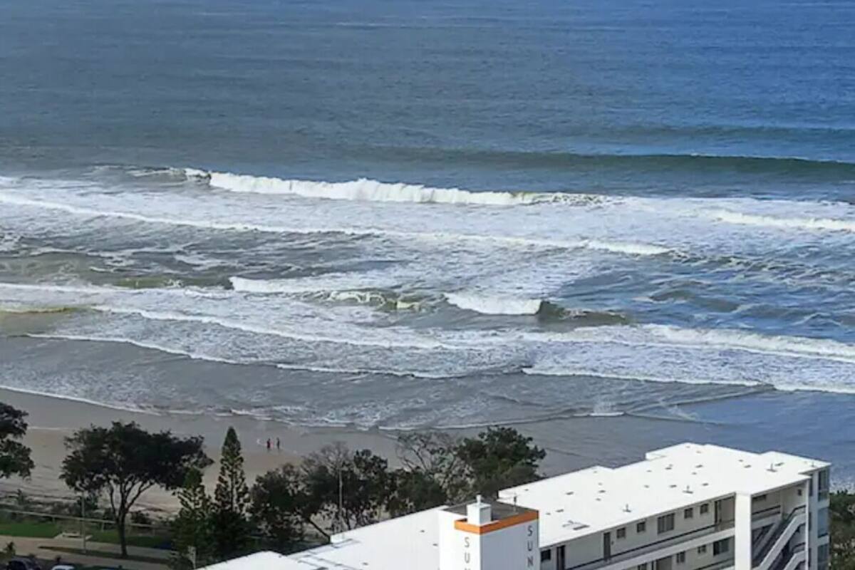 A coastal view with waves crashing onto the shore, a building with a white facade and orange accents, and a clear sky.