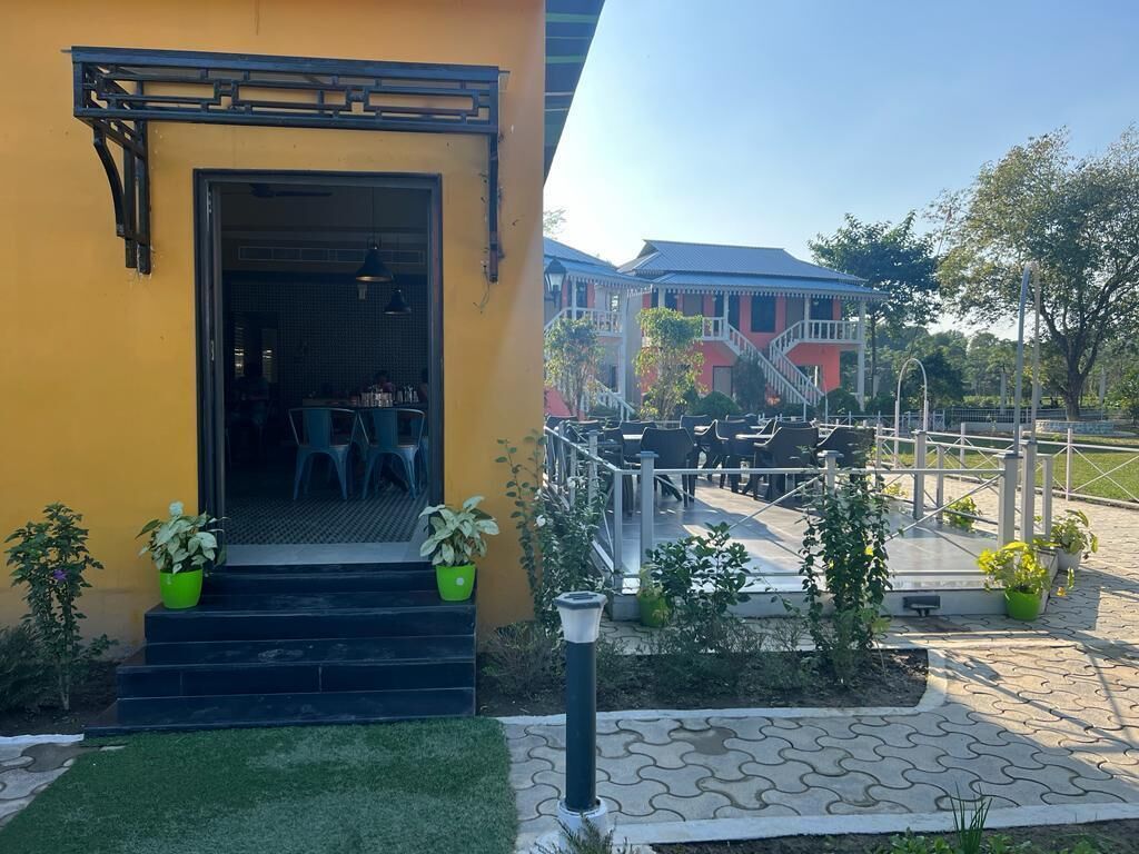 A yellow building with a door leading to a seating area with chairs and tables. There are potted plants on the steps and a paved area with chairs and tables outside.