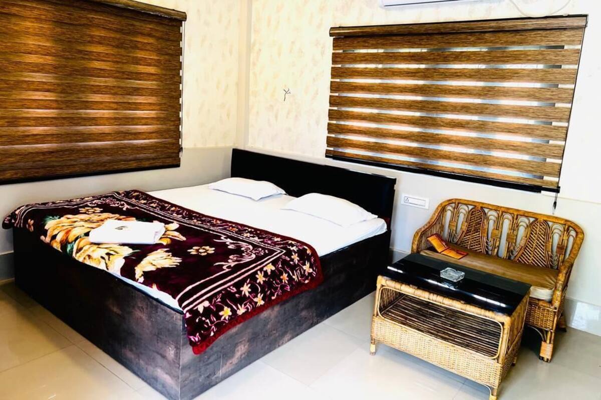A bedroom with a bed, a sofa, and a small table. There are two windows with blinds.