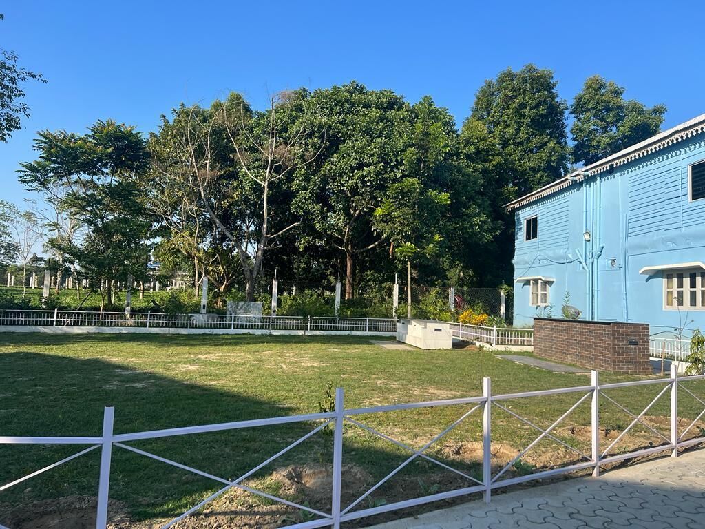 Zona verde cercada con un edificio azul, árboles y cielo despejado.