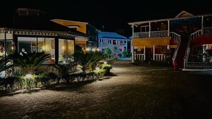Una escena nocturna de un patio empedrado con edificios a ambos lados, destacando una estructura con fachada de vidrio y una escalera que lleva a un balcón en un segundo piso.