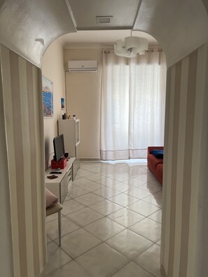 A hotel room with a tiled floor, a desk with a computer, a chair, a bed, a window with curtains, and an air conditioning unit.