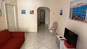 A living room with a red sofa, a TV on a white cabinet, a painting of a coastal scene, and a door leading to another room. - The window in front of Naples (Napoli)