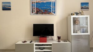 A modern living room with a flat-screen TV on a white cabinet, a small table with a remote and a decorative item, and a cabinet with glass doors displaying bottles and a vase. - The window in front of Naples (Napoli)