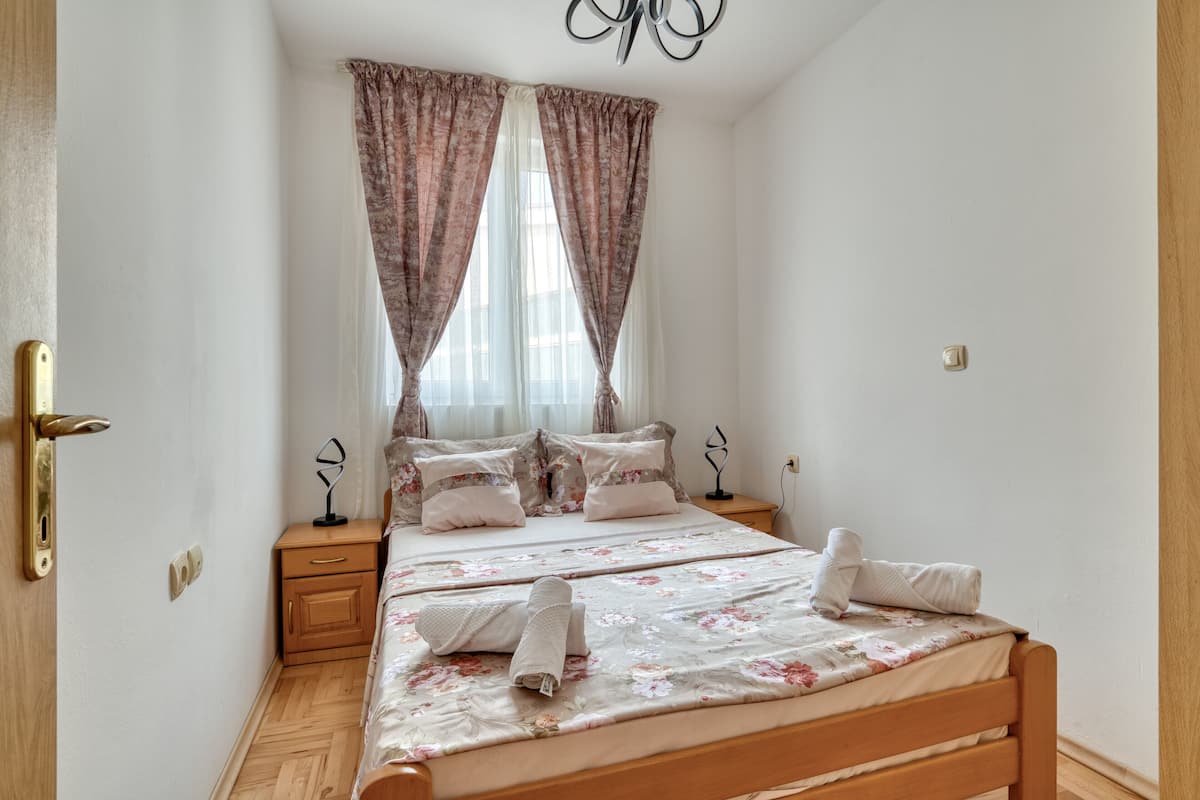 A bedroom with a wooden bed, floral bedding, a nightstand, and a window with curtains.