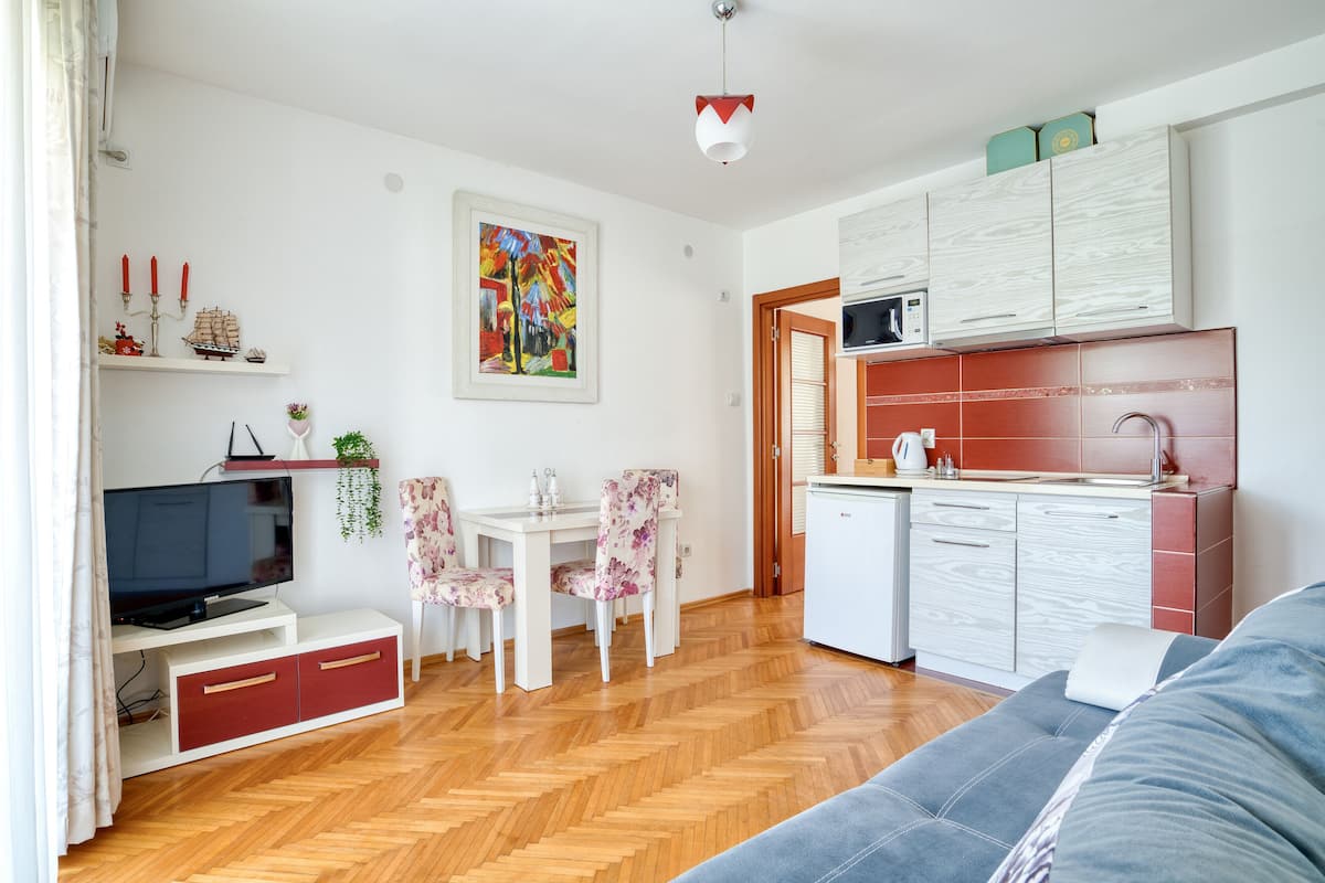 A modern living room with a sofa, TV, dining table, and chairs. A kitchen with a microwave and cabinets is visible.