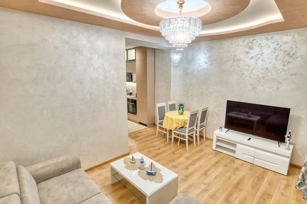 A modern living room with a flat-screen TV, a dining area with a yellow tablecloth, and a chandelier.