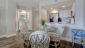 A dining area with a glass table and wicker chairs, a kitchen with stainless steel appliances, and a view of the ocean through a window. - 2 bedroom,  modern oceanfront condo (North Topsail Beach)