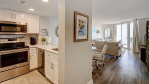 A modern kitchen with stainless steel appliances, a dining area with a white table and wicker chairs, and a living room with a sofa and large windows. - 2 bedroom,  modern oceanfront condo (North Topsail Beach)