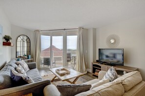 A living room with a sofa, a coffee table, a TV, and a balcony with a view of a building. - 2 bedroom,  modern oceanfront condo (North Topsail Beach)