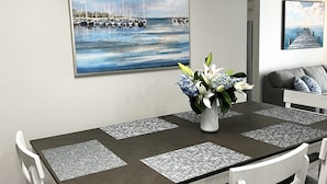 Uma área de jantar com mesa de madeira escura, cadeiras brancas e um vaso com flores. Há duas pinturas na parede, uma retratando uma marina e outra um cais.