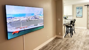 Uma sala de estar com uma grande TV de tela plana na parede, uma área de jantar com mesa e cadeiras, e um ventilador de teto.
