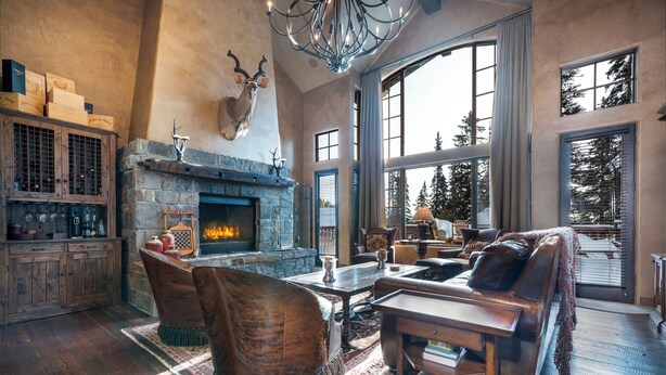 Amplia sala de estar con chimenea de piedra, muebles de cuero y grandes ventanales con vista a árboles.
