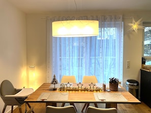 A dining area with a wooden table, chairs, a hanging light fixture, and a window with sheer curtains.