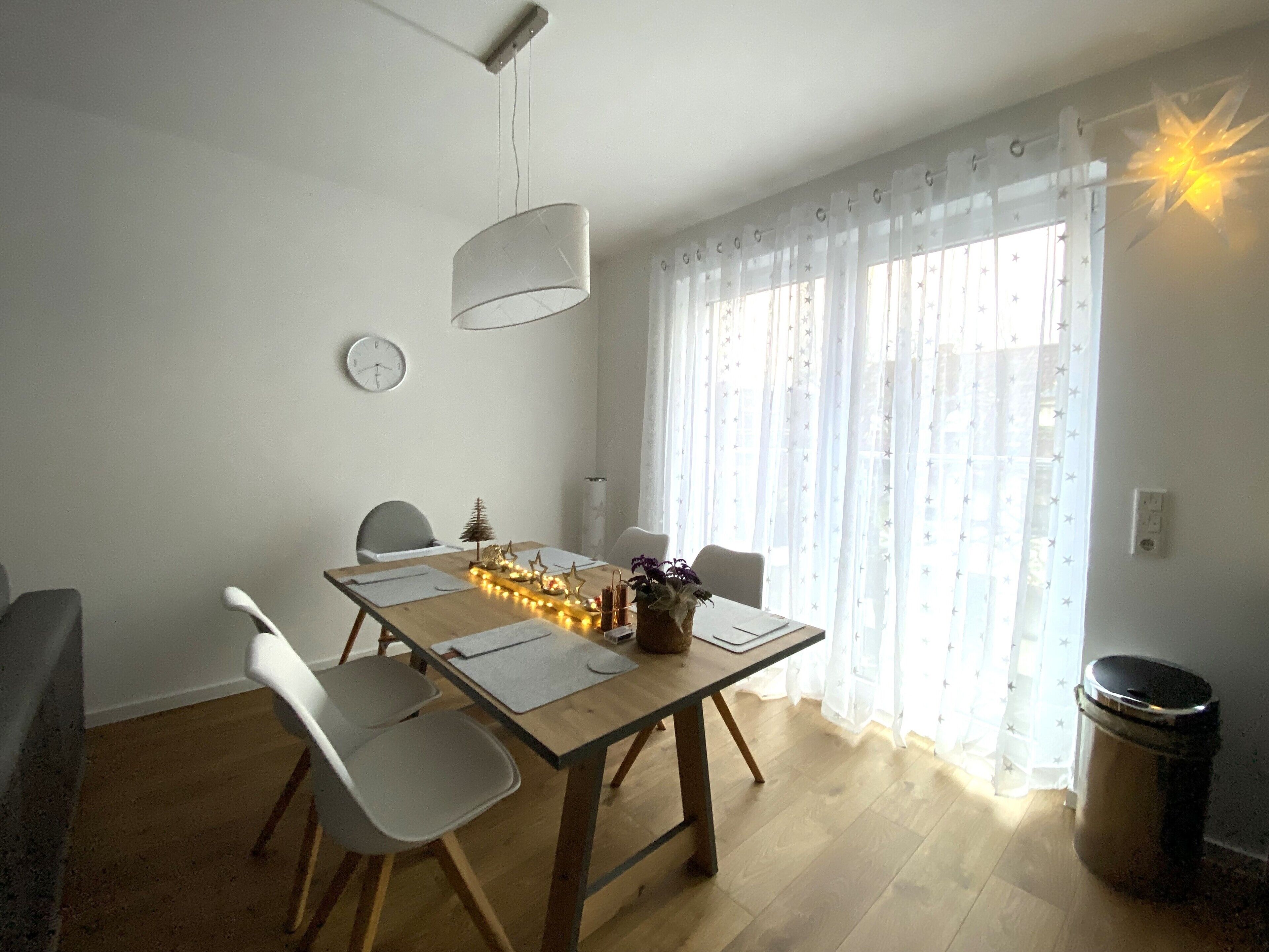 A dining area with a wooden table, white chairs, a hanging light fixture, a small Christmas tree, and a star-shaped decoration.