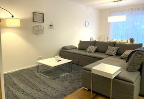 A modern living room with a grey sofa, two white side tables, a grey shag rug, and a large window with sheer curtains.