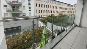 A balcony with a glass railing overlooking a garden with orange flowers and a building with multiple windows in the background. - Mein Apartment, Stars for 4 Person (Frankenthal)