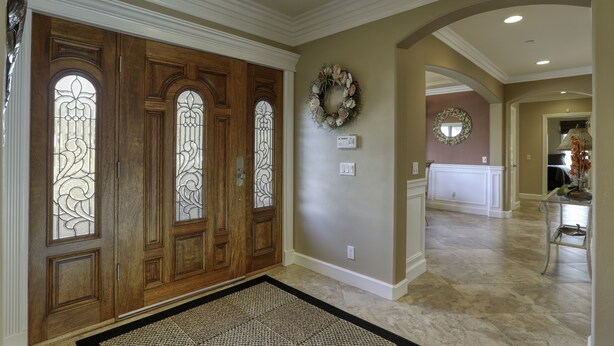 A spacious hallway with a wooden door with stained glass panels, a decorative wreath on the wall, and a tiled floor.