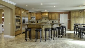 A spacious kitchen with wooden cabinets, a large island with bar stools, stainless steel appliances, and a tiled floor.