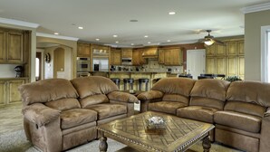 A spacious living room with leather sofas and a coffee table, a kitchen with wooden cabinets and modern appliances, and a ceiling fan.