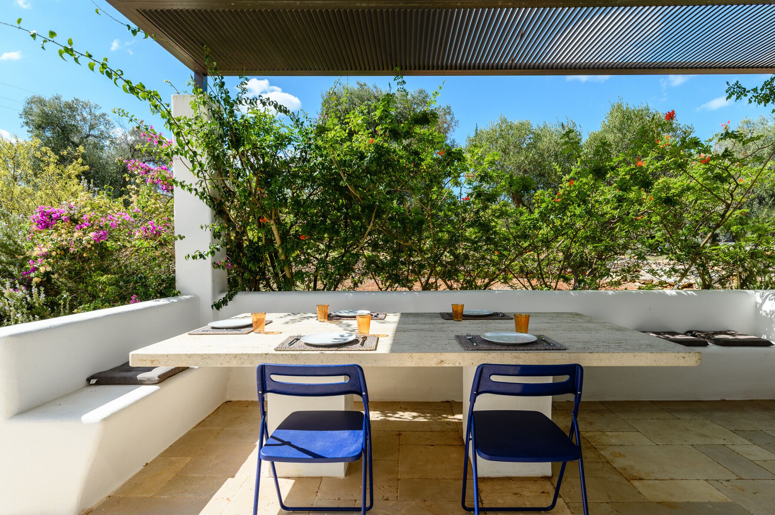 Un espace de restauration en plein air avec une table dressée pour deux personnes, entourée de chaises, sous une pergola offrant une vue sur une végétation luxuriante et des arbres.
