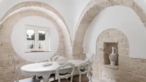 A dining area with a round table, white chairs, and a stone wall with a window. - Trulli Julia con Piscina by Wonderful Italy (Martina Franca)