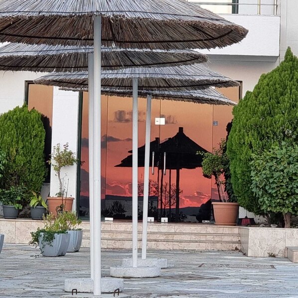 A building entrance with a large umbrella structure, potted plants, and a view of a sunset over a mountain. - Hotel Mercury Saranda (Sarandë)