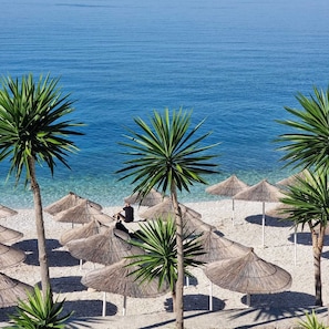 A beach with palm trees and thatched umbrellas. - Hotel Mercury Saranda (Sarandë)