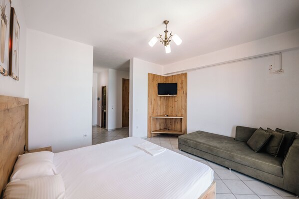 A modern bedroom with a bed, a wooden headboard, a grey sofa, and a mounted TV. - Hotel Mercury Saranda (Sarandë)