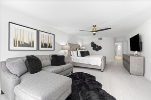 A modern bedroom with a grey sofa, a bed with a white headboard, a black furry rug, and a ceiling fan. - Upscale Oceanfront – Club Longboat #334 RVA (Longboat Key)