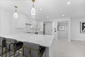 A modern kitchen with a large island, bar stools, pendant lighting, and white cabinetry. - Upscale Oceanfront – Club Longboat #334 RVA (Longboat Key)