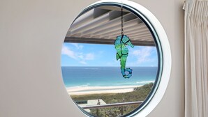 Une fenêtre ronde donnant sur une plage et l’océan.