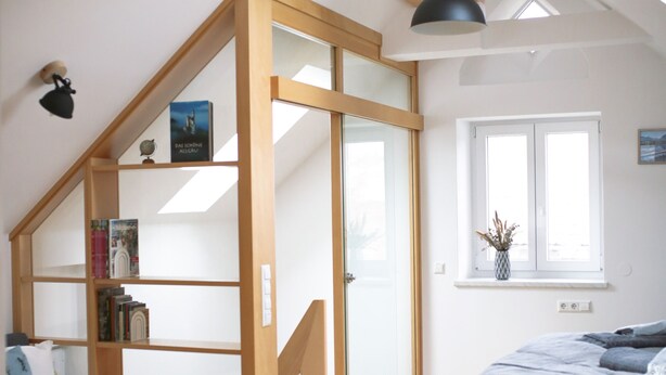 Une chambre à coucher aménagée dans un espace en hauteur, avec un escalier en bois, un lit, une bibliothèque et une fenêtre donnant sur un paysage.