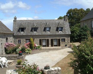 A stone building with a gravel courtyard, surrounded by flowering plants and trees. - Maison de Campagne (Plonévez-Porzay)