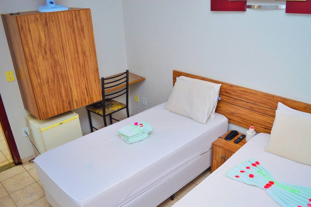 A single bed with a wooden headboard, a bedside table with a phone and remote, a chair, and a wooden wardrobe.