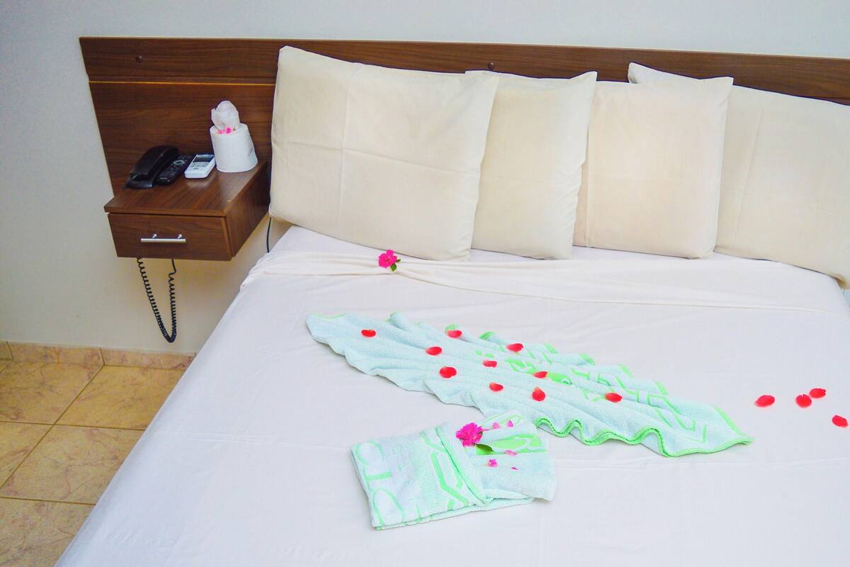 A hotel room with a bed, pillows, a bedside table with a phone, and a nightstand.