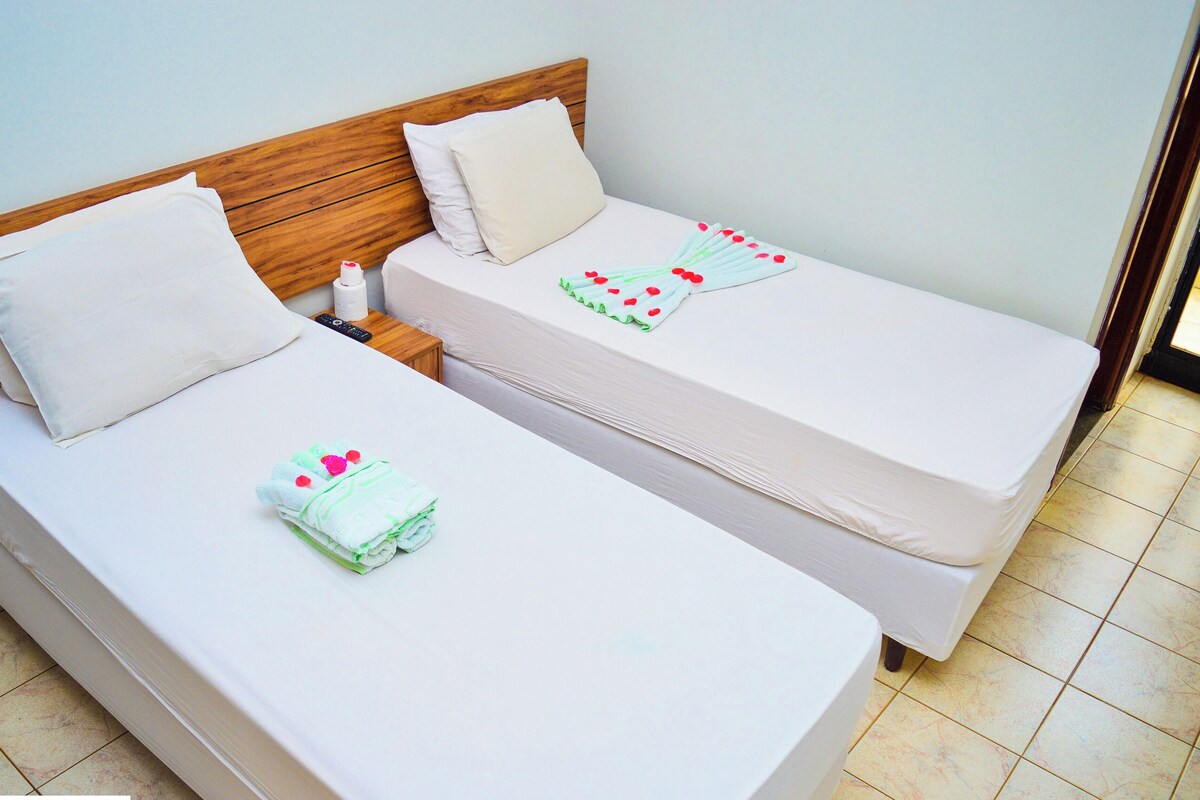 Deux lits simples avec des têtes de lit en bois, du linge de lit blanc et une serviette verte agrémentée de touches de rouge.