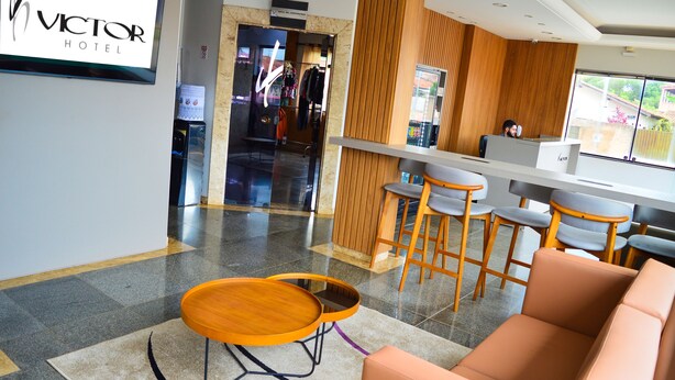 A modern hotel lobby with a reception desk, bar area, seating, and a visible clothing rack. - Victor Hotel (Patos de Minas)