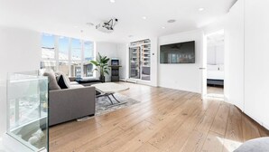 Een moderne woonkamer met een bank, een salontafel en een televisie. Er is een groot raam met uitzicht op gebouwen en een balkon.