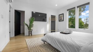 A modern bedroom with a large bed, a flat-screen TV, a window with a view of trees, and a potted plant. - Modern Home with Rooftop Deck (Atlanta)
