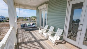 Une véranda couverte avec une terrasse en bois, des chaises blanches et un banc rembourré agrémenté de coussins.