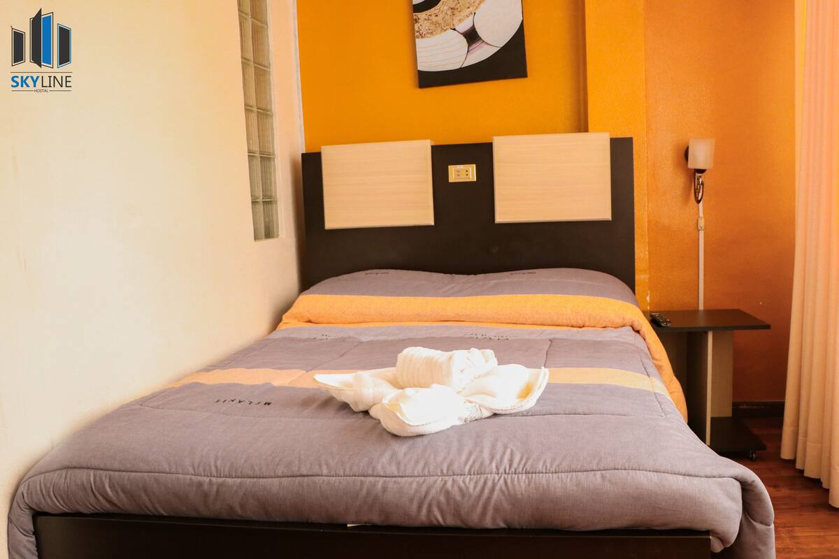 Ein ordentlich bezogenes Bett mit einer grau-orangenen Tagesdecke, einem schwarzen Kopfteil und einem an der Wand hängenden Kunstwerk darüber.
