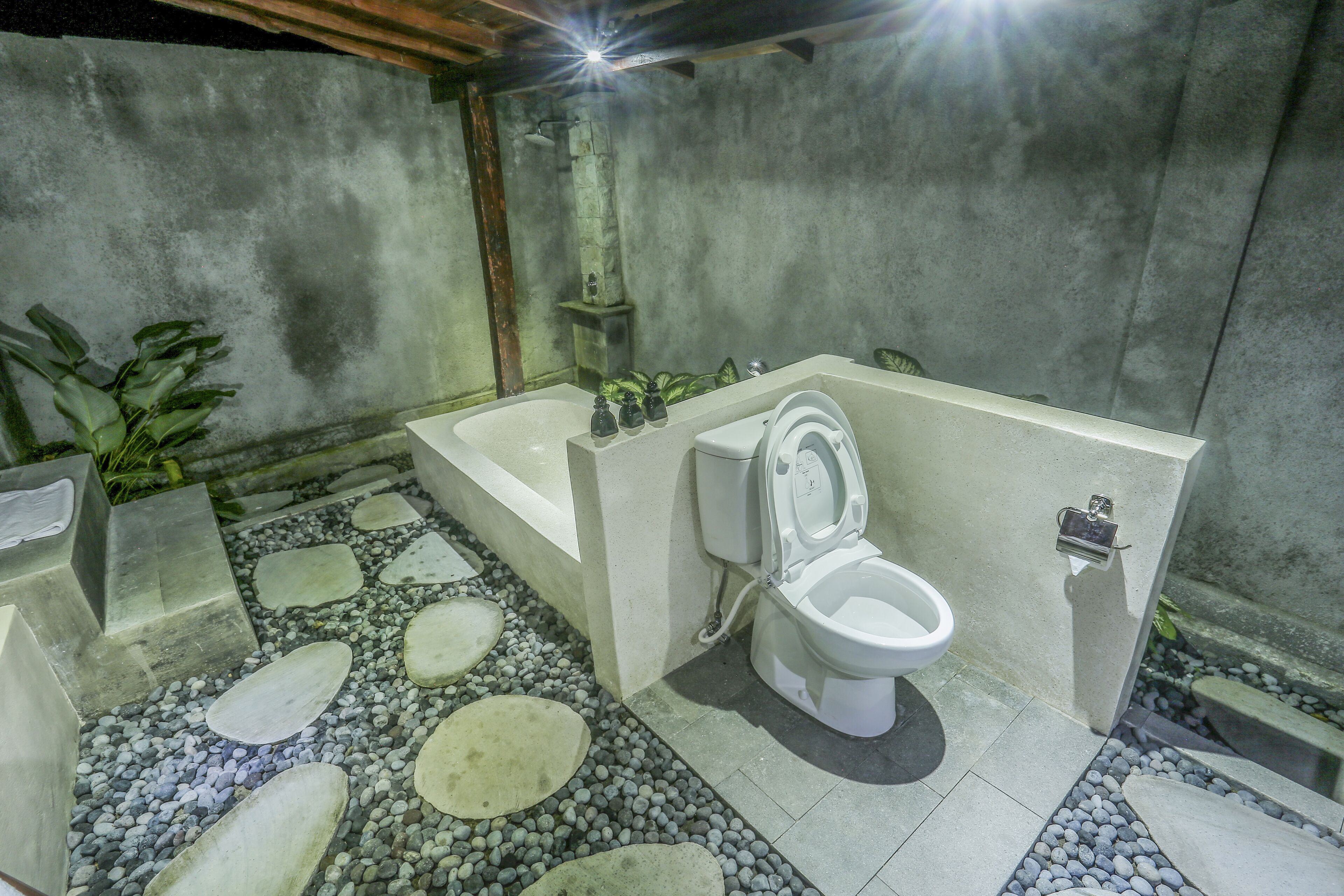 Une salle de bain avec une baignoire, des toilettes et un petit lavabo. Le sol est recouvert de grandes carreaux blancs, entrecoupés de petits galets.