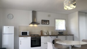 Eine moderne Küche mit weißen Schränken, einem eingebauten Backofen, einer Mikrowelle, einem Gasherd und einem Spülbecken. Es gibt einen Esstisch mit Stühlen, eine Uhr an der Wand und ein Fenster mit Blick ins Grüne.
