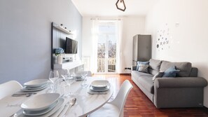 Una sala de estar moderna con un sofá gris, una mesa de comedor con platos y vasos, un televisor montado en la pared y un ventanal con vistas a edificios.