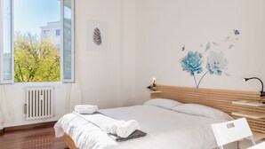 Een slaapkamer met een bed, een radiator, een raam met uitzicht op bomen, een muur met een dennenappel en bloemen, en een bureau met een lamp.