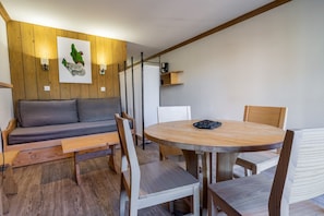 Um quarto acolhedor com parede de madeira, um sofá cinzento, uma mesa redonda de madeira com cadeiras e uma máquina de café.